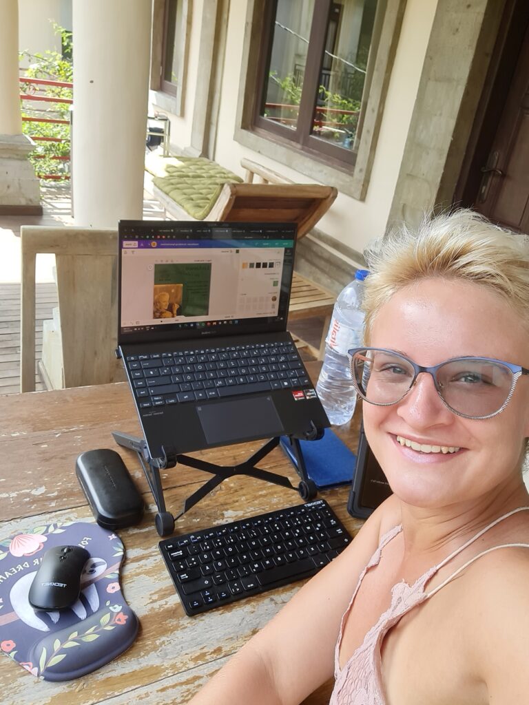 A blonde woman turning over her shoulder to look up from her outside office work station. Her laptop is mounted on a stand and one can see a black laptop mouse on the left and a remote keyboard in front of the laptop.She is wearing a pink top.