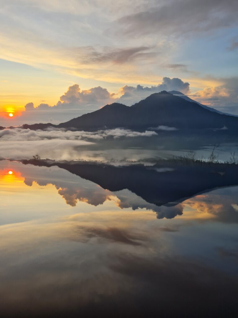 two mount volcano that is reflected in the lake below. Sun rises in the far left back and the tip of the volcano is covered in clouds.