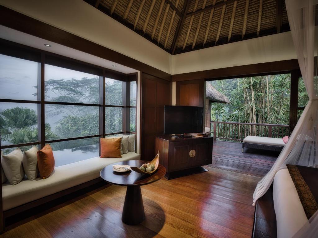 Lush green nature surrounds this private villa in Ubud with a private pool. One can see a couch on the left, which overlooks the pool in the background through large windows. there is a coffee table in front of the couch, a TV towards the back, and once can see the corner of the king side bed on the right. 