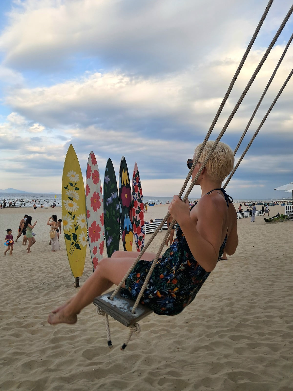 location independent coaching businesses: a woman with a short dress and short blonde hair sitting on a swings looking out over the ocean. there are surfboards stacked in the sand behind her.