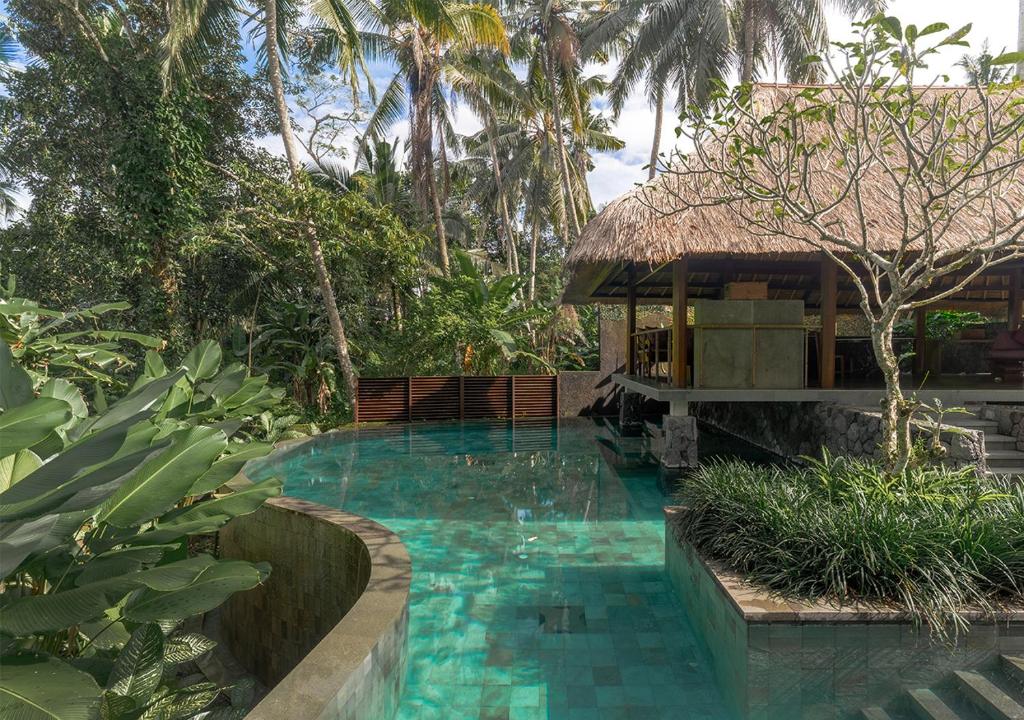 The Katumanis Ubud private villas might be amongst the best hotels in Ubud with private pools for working digital nomads. A large infinity pool with turquoise water is shown, surrounded by green nature. A bamboo covered structure is in the background which hints at the entrance into the private villa. 