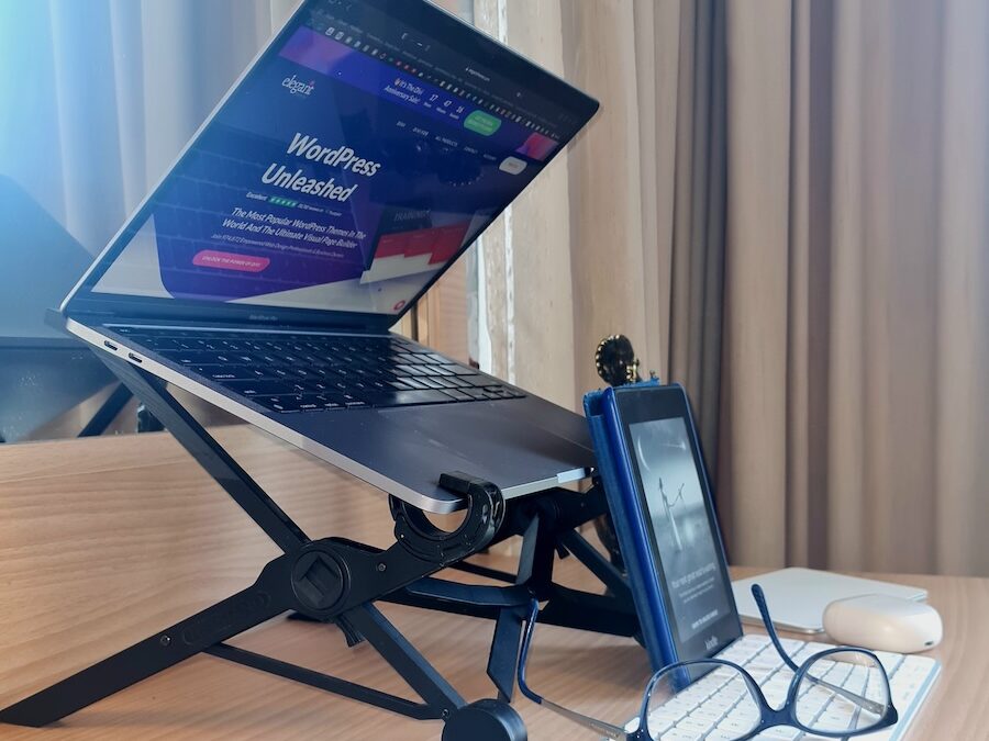 a grew Macbook laptop is shown on a tilted laptop stand that is black. A blue box is in front, a blue pair of vision glasses rests across a white magic keyboard. A blue and black Kindle rests against the laptop stand, right behind the keyboard.