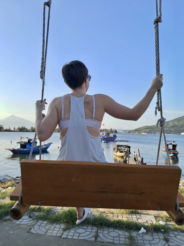 woman who is known as creator of Way of the Founder and business and mindset coach for location independent entrepreneurs, sits on a swing set with her back to the camera, overlooking a river, with mountains in the background. she's wearing a white tank top and has dark blonde, short hair.